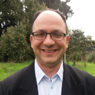 A man smiling, wearing glasses outdoors