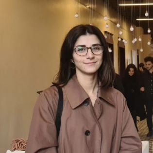 A woman wearing glasses indoors, standing in front of a wall.