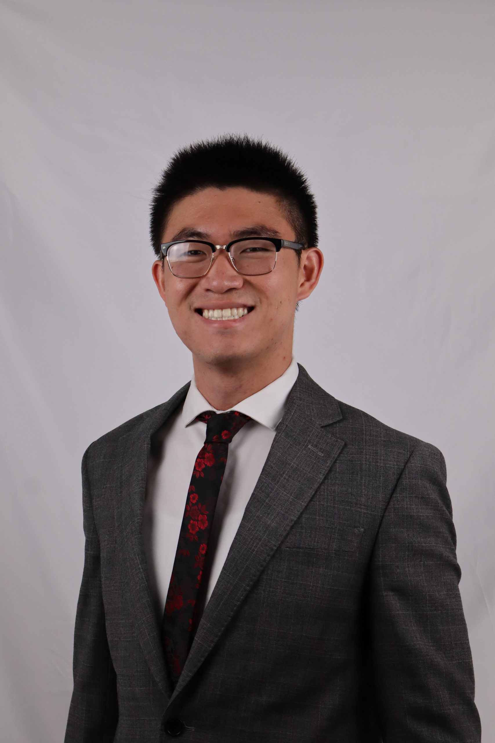 Smiling person wearing glasses, a grey suit jacket, and a dark patterned tie